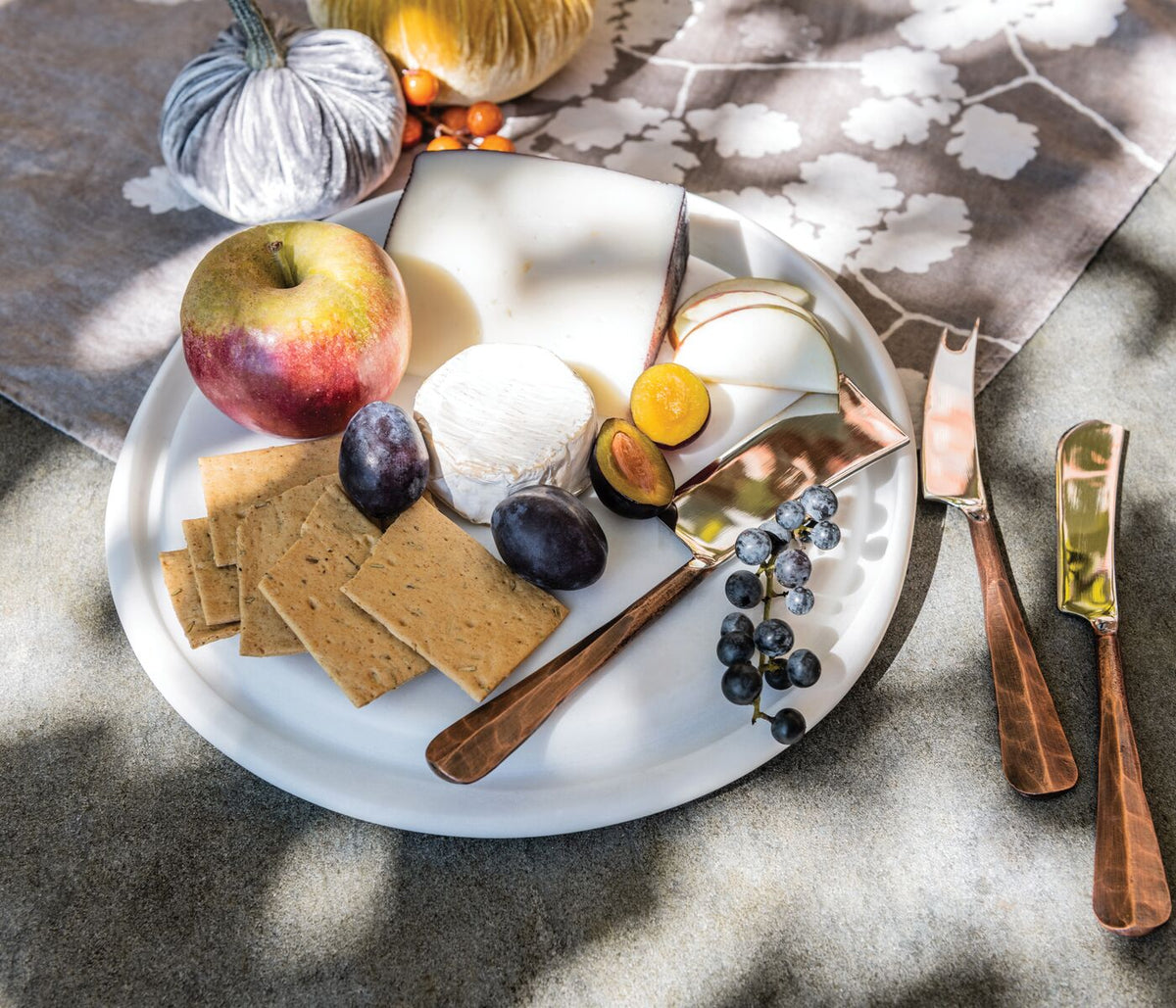 White marble round board filled with cheese, crackers, fruit, and wooden spreader knife.