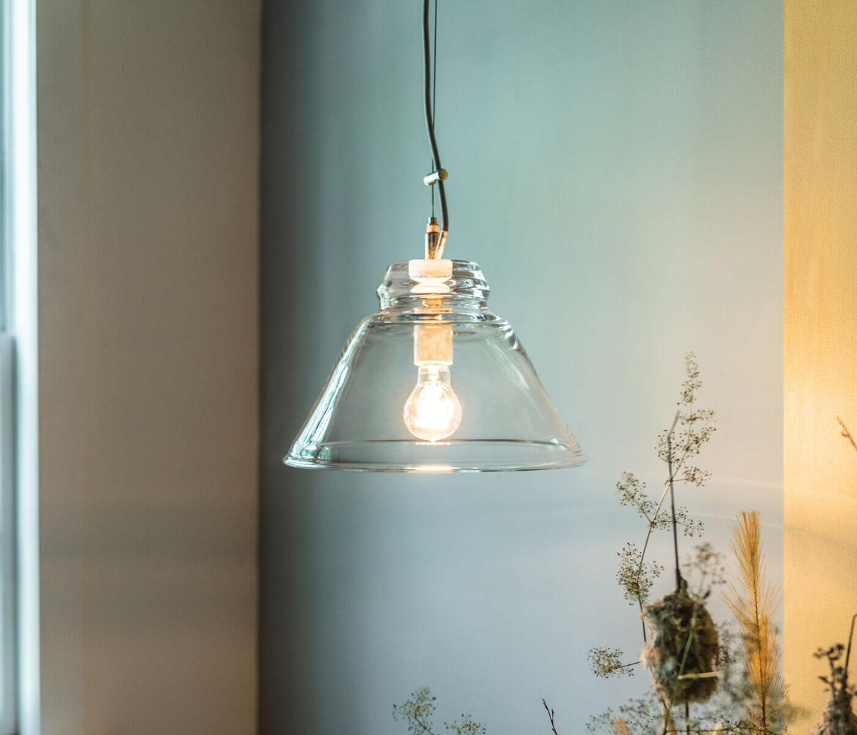 Clear glass cone-shaped pendant with brass fitting and black cord, lit incandescent bulb visible inside.