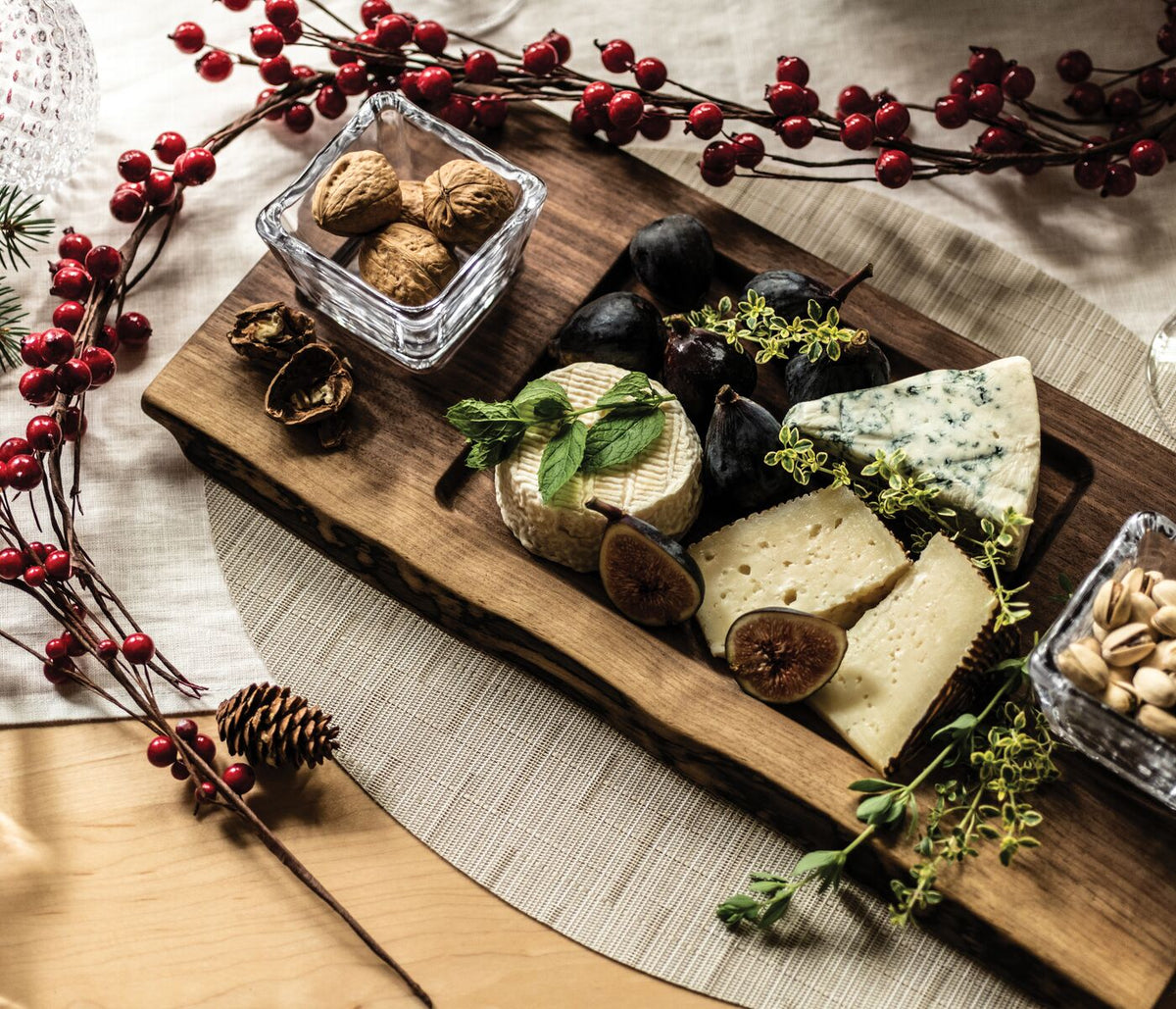 Black walnut serving board with glass bowl filled with walnuts, arranged with cheese, herbs, and dried berries.