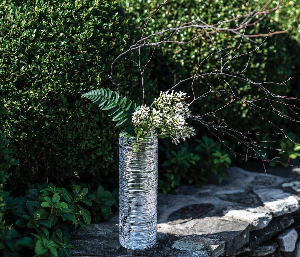 Clear glass cylindrical vase with horizontal ridged texture containing white flowers and green fern stems.