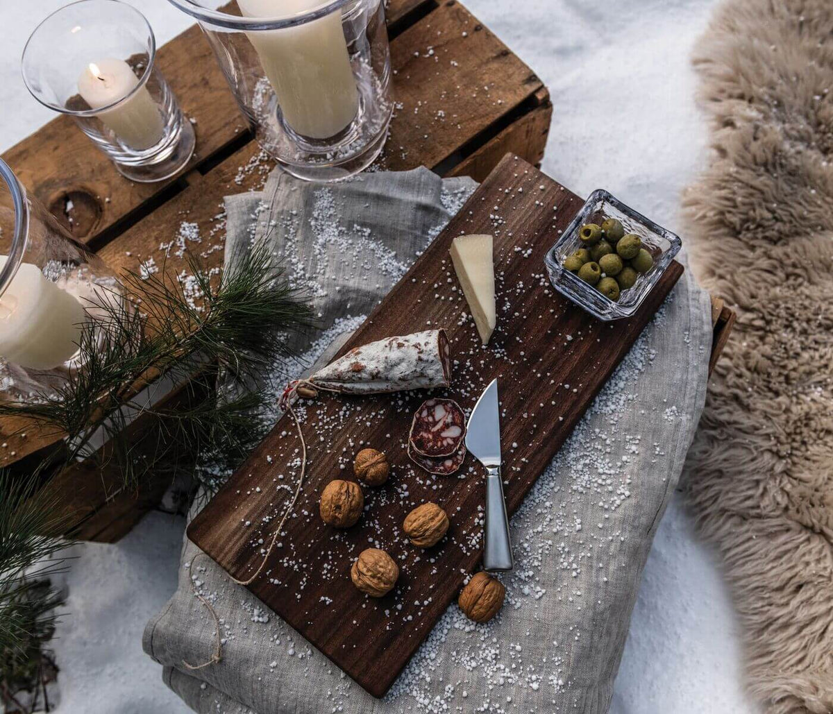 Dark walnut live edge board displaying cheese, olives, walnuts, and a knife, with glass bowls and candles on gray linen.