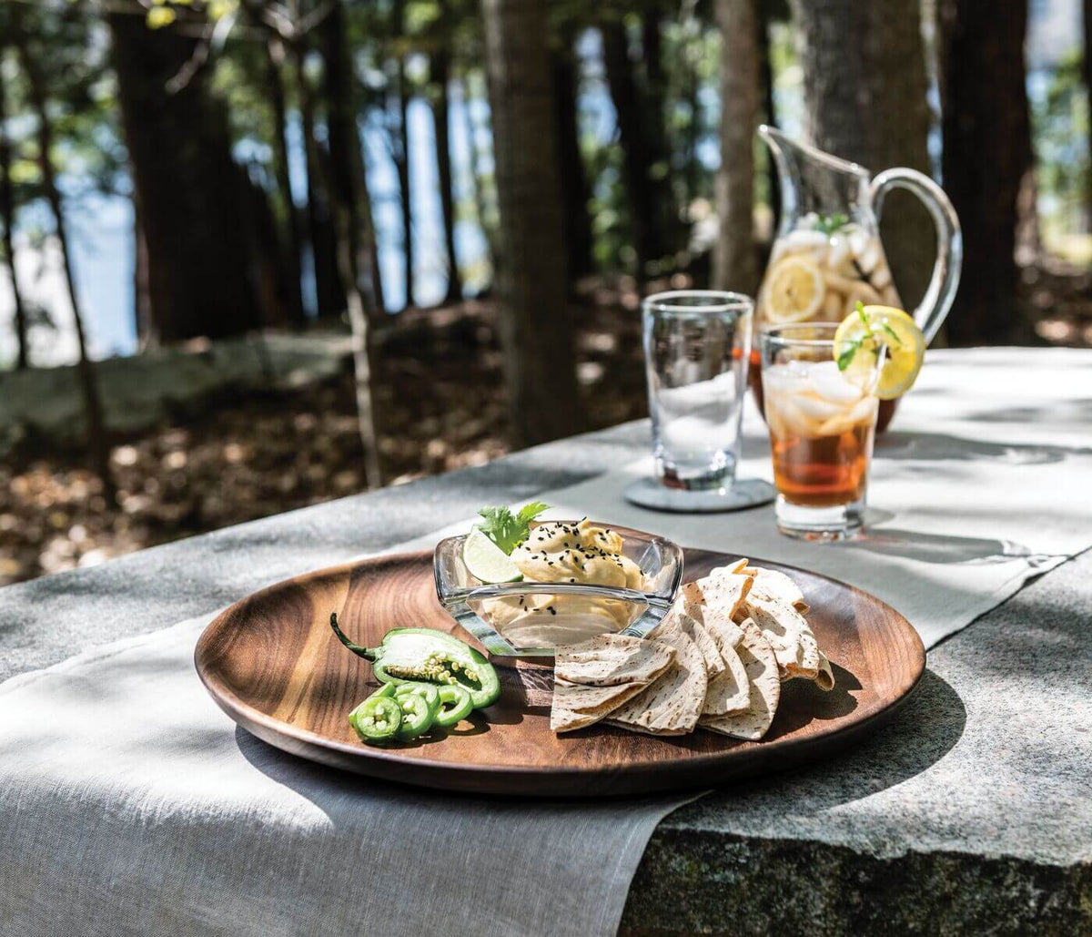 Wooden serving platter with glass dip bowl holding white dip, surrounded by tortilla chips and sliced jalapeños.
