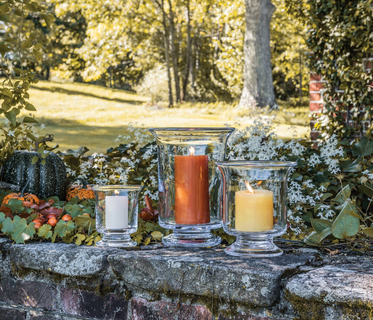 Three glass Hurricane candles in varying heights with white, amber, and cream-colored wax displayed outdoors on a stone surfa