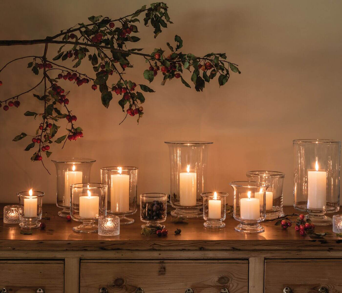 Multiple clear glass hurricane candles with lit white pillar candles arranged on wooden dresser with dried berries and foliag