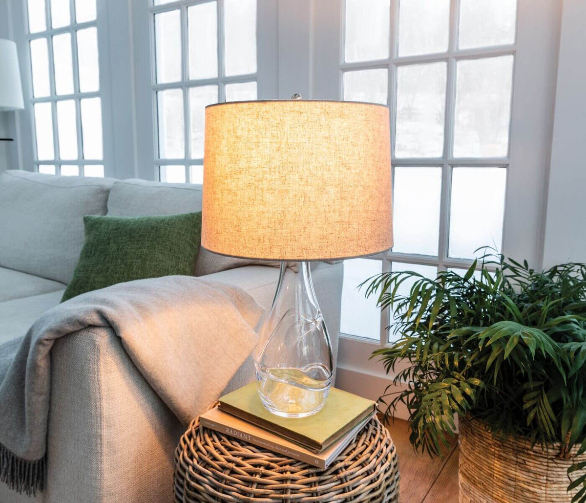Table lamp with clear glass base, tan linen shade, and metal hardware on wicker side table.
