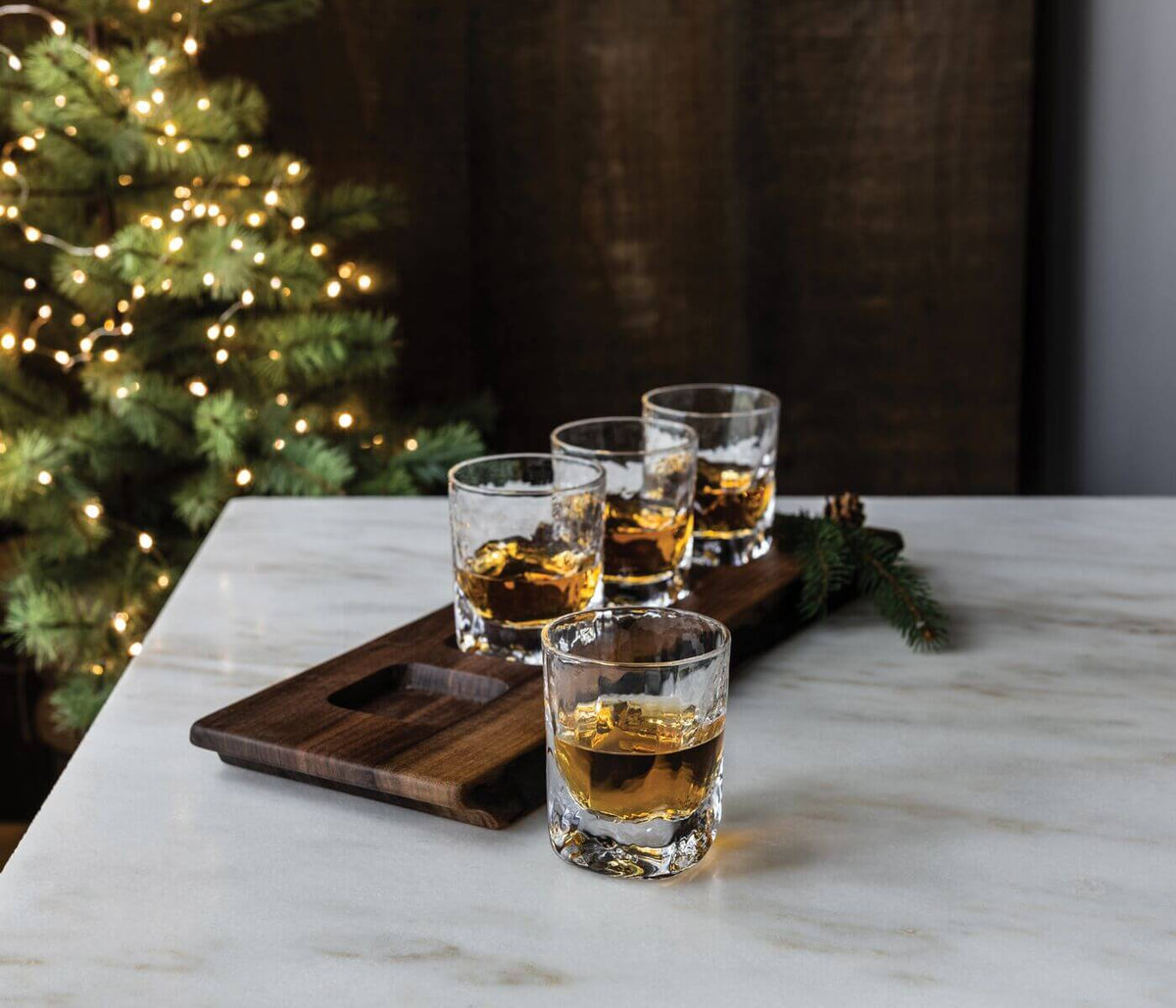 Four glasses filled with bourbon and ice arranged on a dark wood serving board with evergreen garnish.