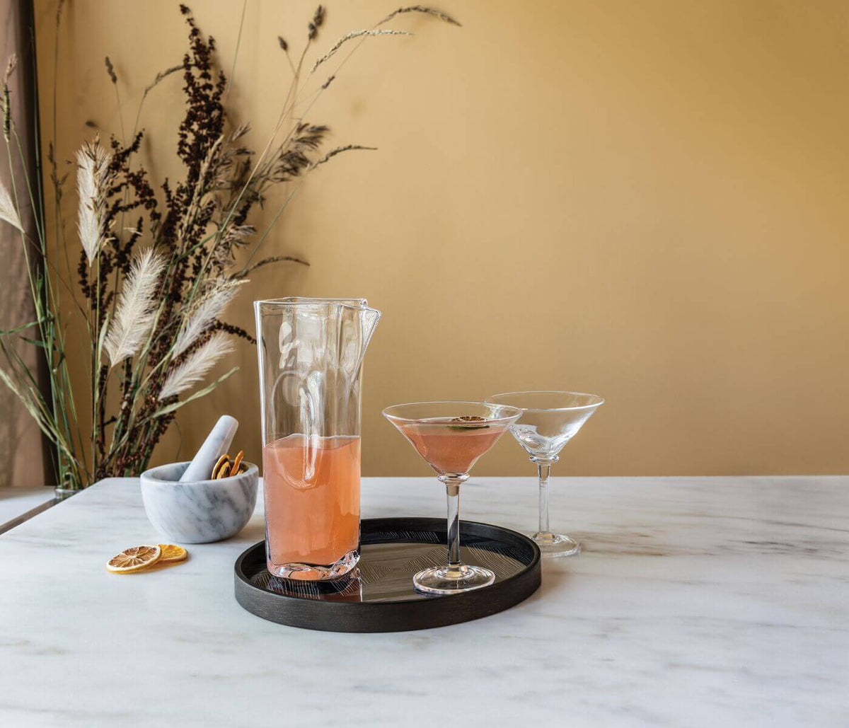 Clear glass carafe filled with coral-colored liquid, displayed on a black tray with two cocktail glasses.