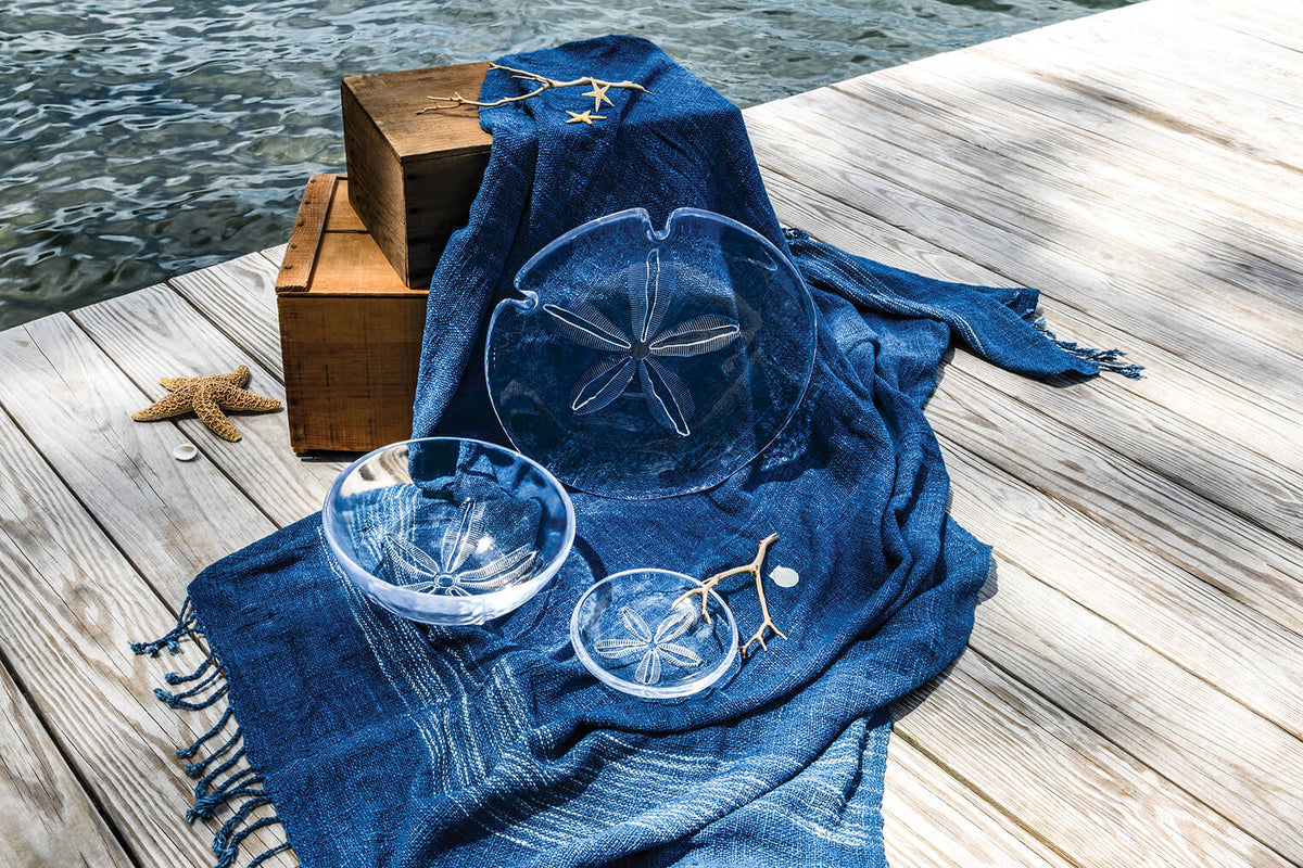White ceramic plate with sand dollar relief design on blue fabric beside wooden boxes on a dock.