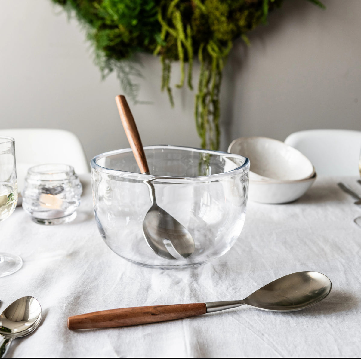 Clear glass bowl with wooden and metal spoon inside, displayed on white table with place settings.