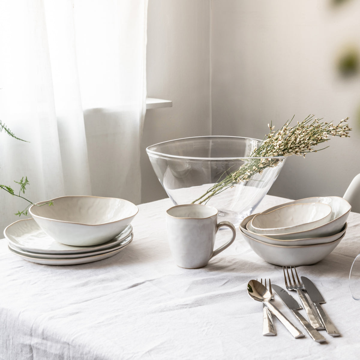 White dinnerware set with bowls, plates, mug, and serving dish arranged on table with cutlery and dried flowers.
