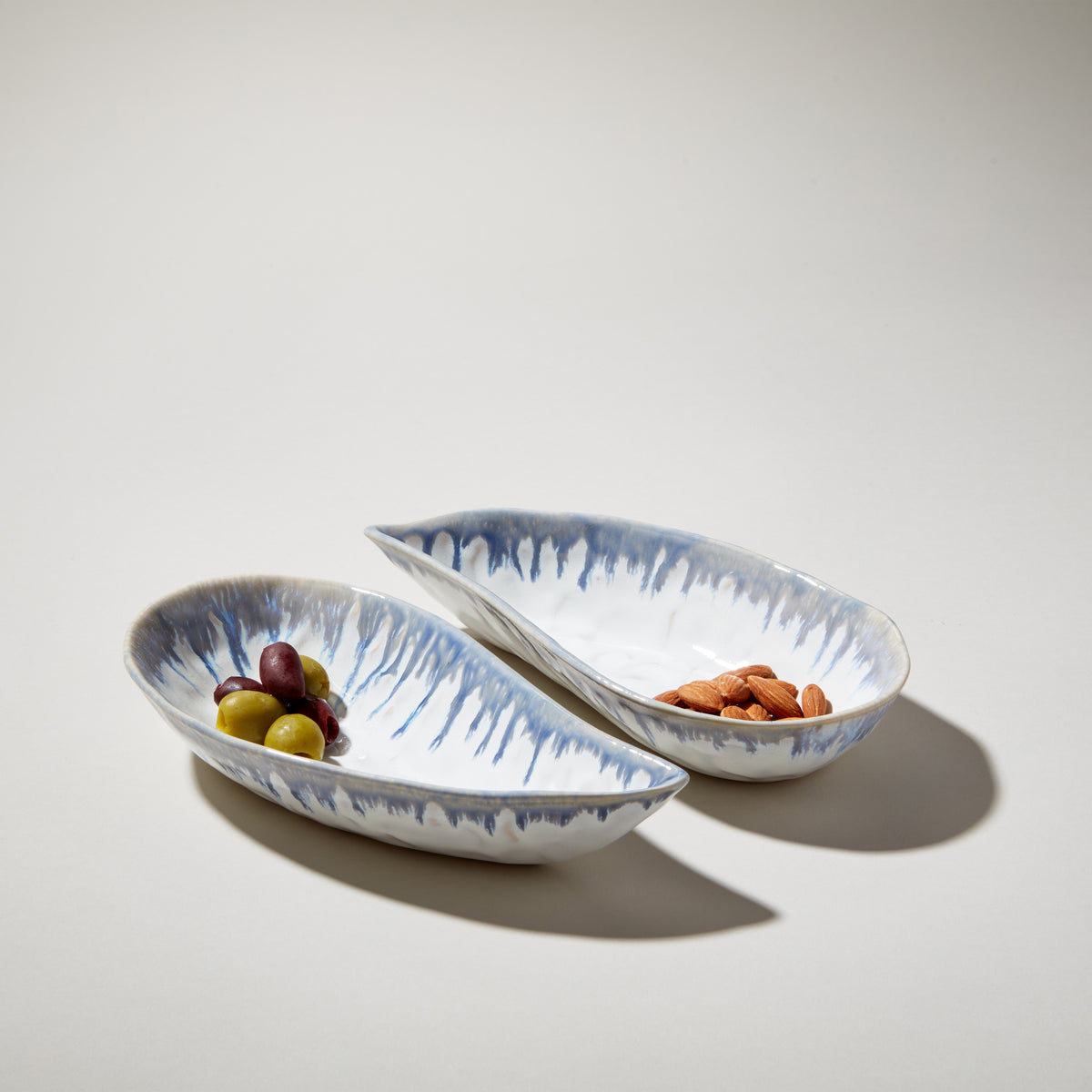 Two white mussel-shaped bowls with blue brush stroke pattern, one containing olives and one containing almonds.