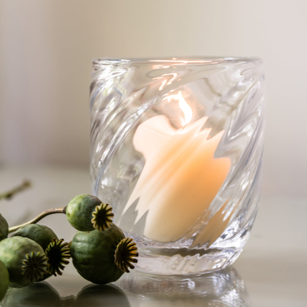 Clear glass candle with cream-colored wax and textured relief pattern, accompanied by green poppy seed pods.