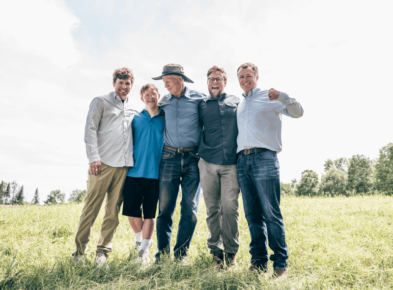 Five men standing together and laughing in a green field, representing multiple generations of the Pearce family