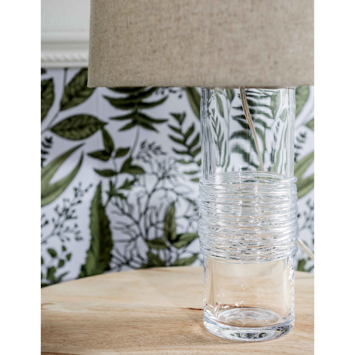 Clear glass cylindrical lamp with textured upper section, linen shade, and botanical leaf patterned pillow base.