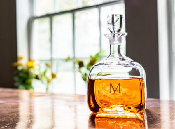 A Simon Pearce glass decanter engraved with the letter M, filled with amber whiskey, on a marble surface