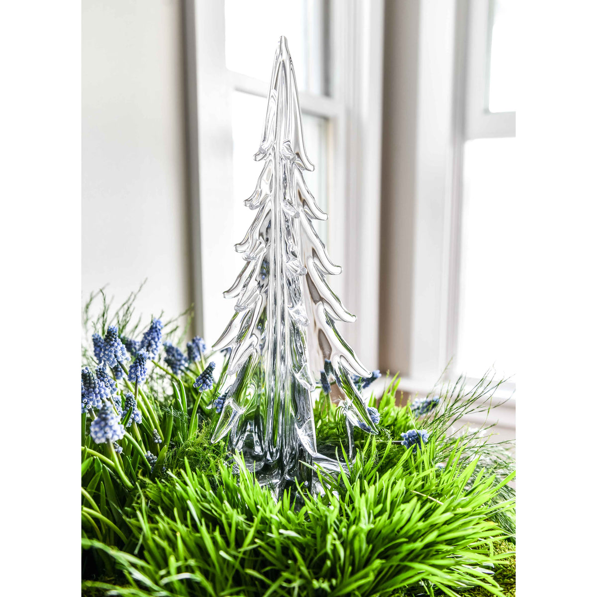 Clear glass evergreen tree with tiered branches displayed among green foliage and blue flowers by a window.