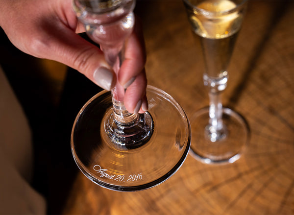 A hand holding the base of a Simon Pearce champagne flute engraved with a wedding date, with a second flute in the background