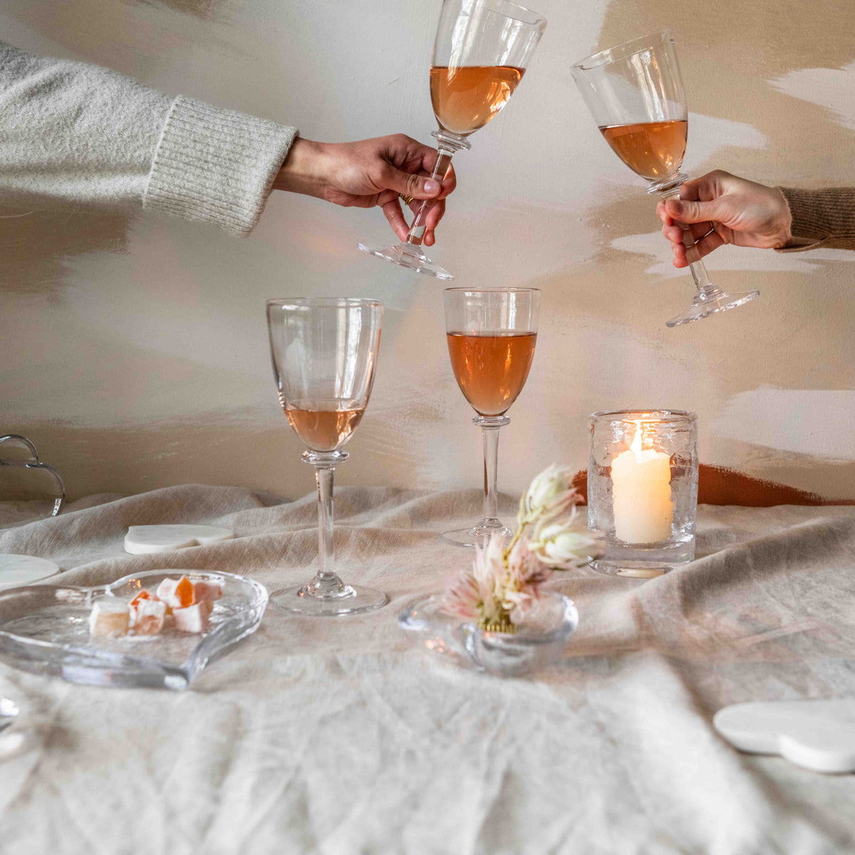 Two hands holding wine glasses filled with amber-colored wine on a neutral-toned table setting.