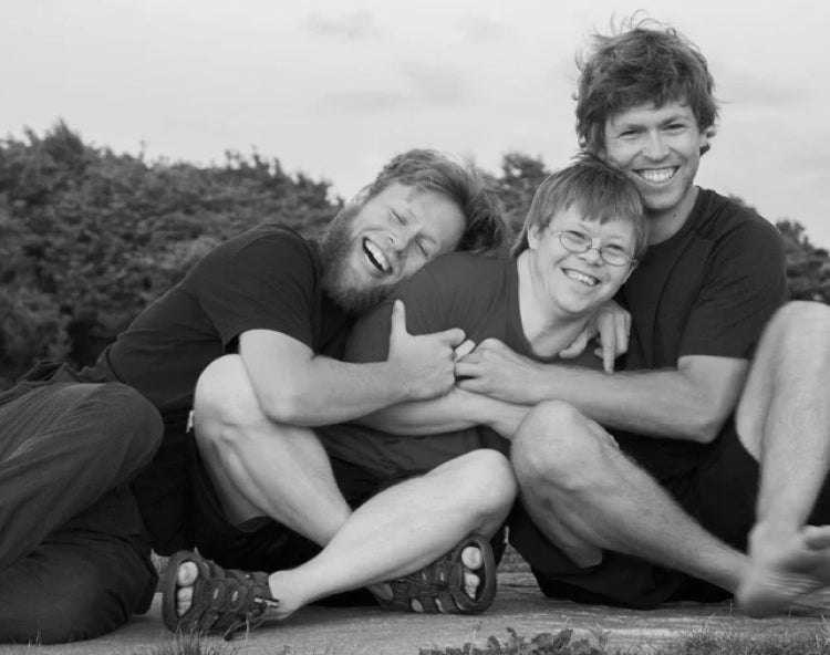 Three people laughing and embracing on a lawn in a black-and-white photo, capturing the joy and connection at the heart of LoveYourBrain