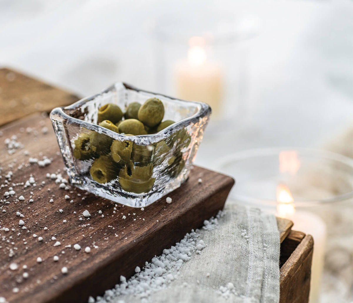 Black walnut live edge board with square glass bowl containing green olives, styled with sea salt and candles.