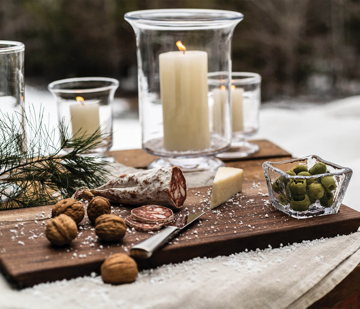 Dark wood board with live edge holding walnuts, cured meat, cheese, and greens next to glass bowl and lit candles in glass ho