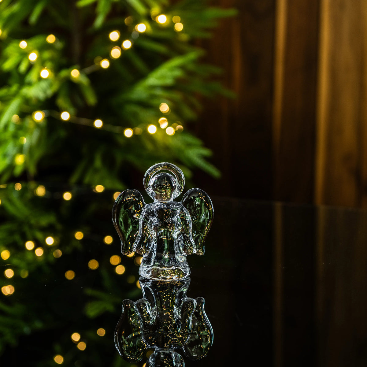 Small glass angel figurine with halo and wings, displayed with evergreen garland and warm string lights.