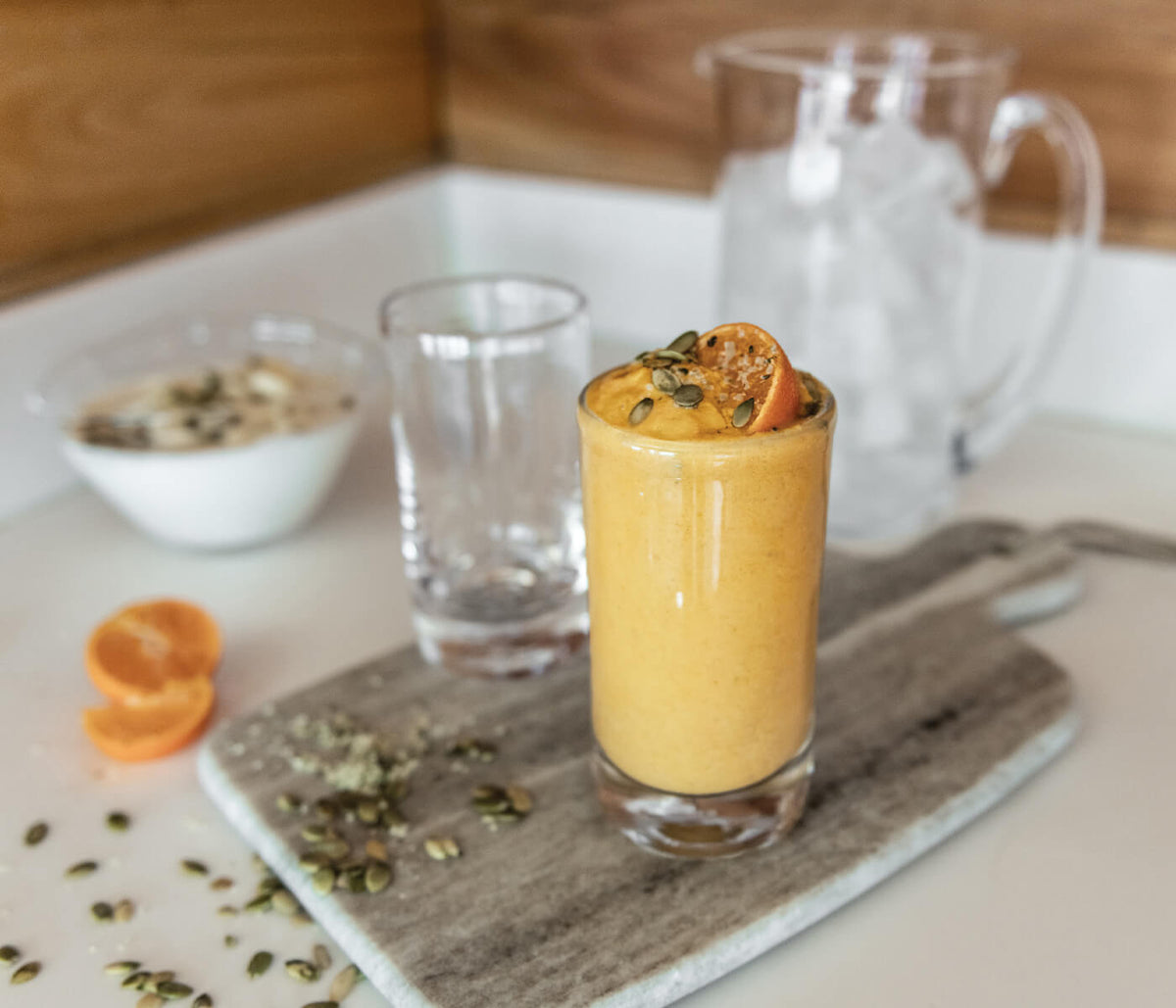 Two clear highball glasses with one filled with yellow cocktail and orange slice garnish, displayed with pitcher and table se