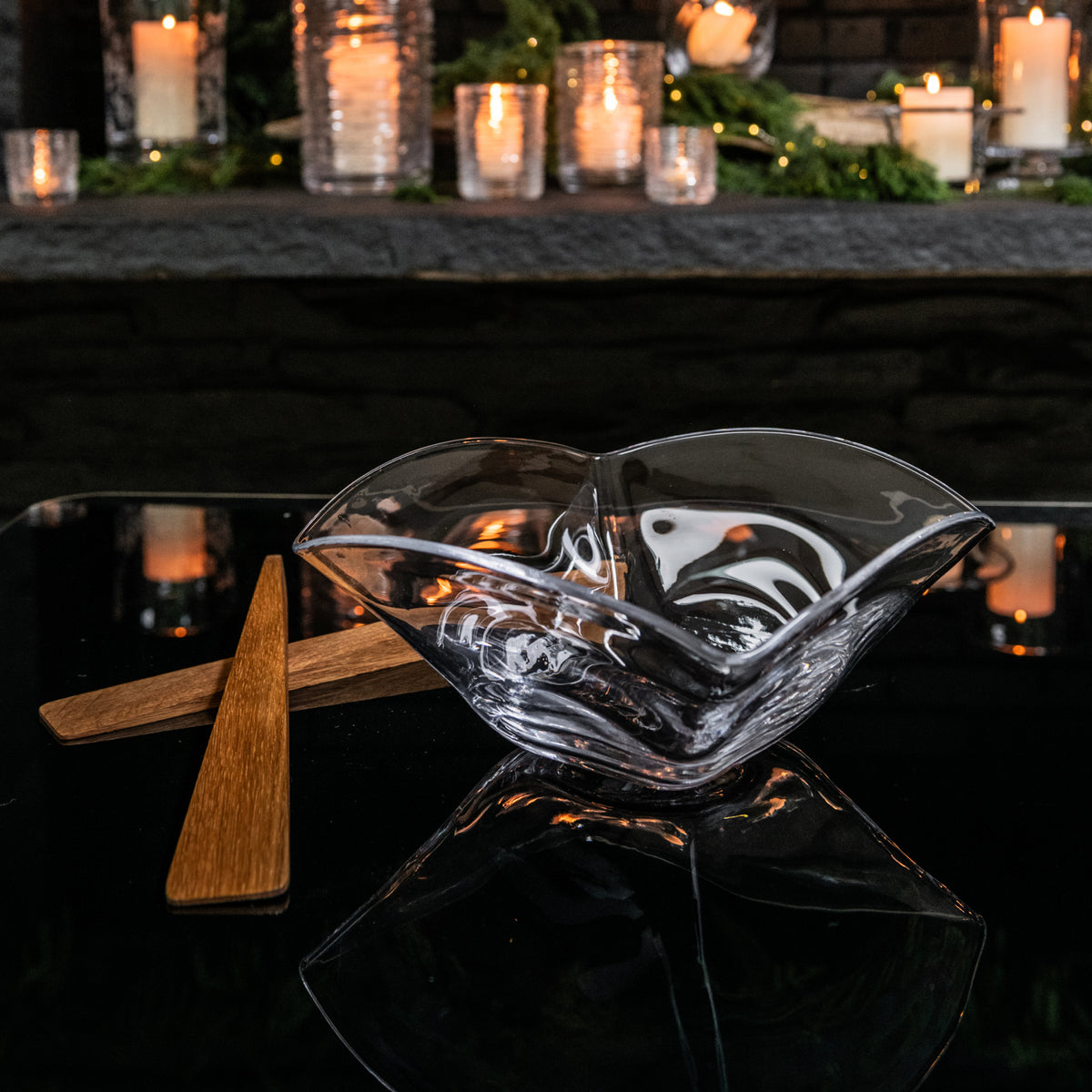 Clear glass serving bowls with wooden handles on a dark surface, surrounded by lit candles and greenery.