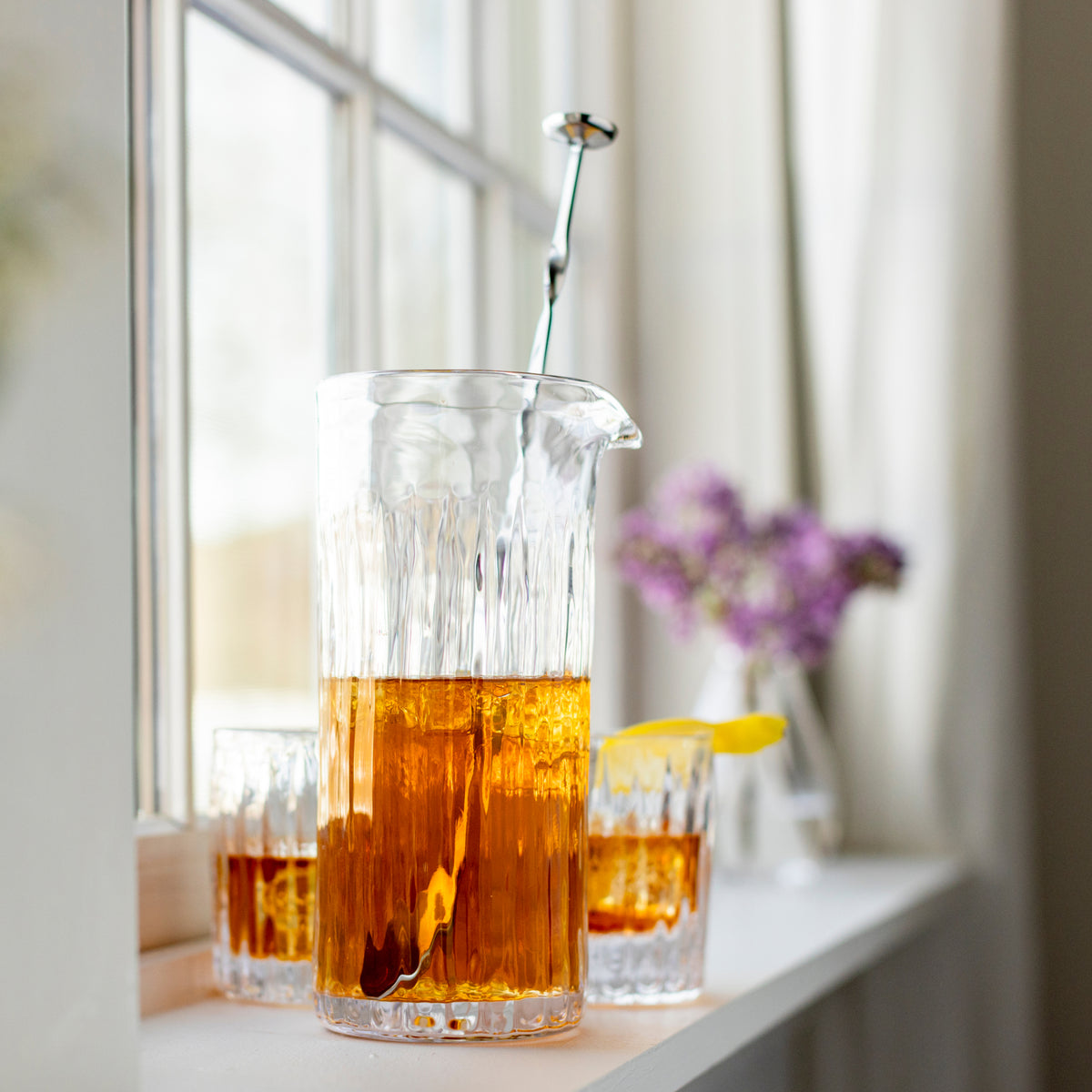 Stainless steel stirring spoon with long handle and small flat bowl head resting in a glass pitcher of amber liquid.