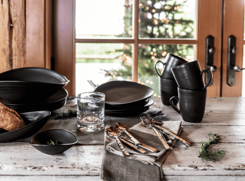 A rustic wooden table set with matte black stoneware plates, bowls, and mugs alongside a clear drinking glass and copper-handled flatware, with a lit Christmas tree visible through the window behind