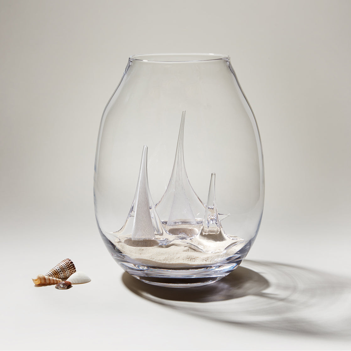 Clear glass globe with three tall sailboat figurines and sand inside, displayed with small shells.