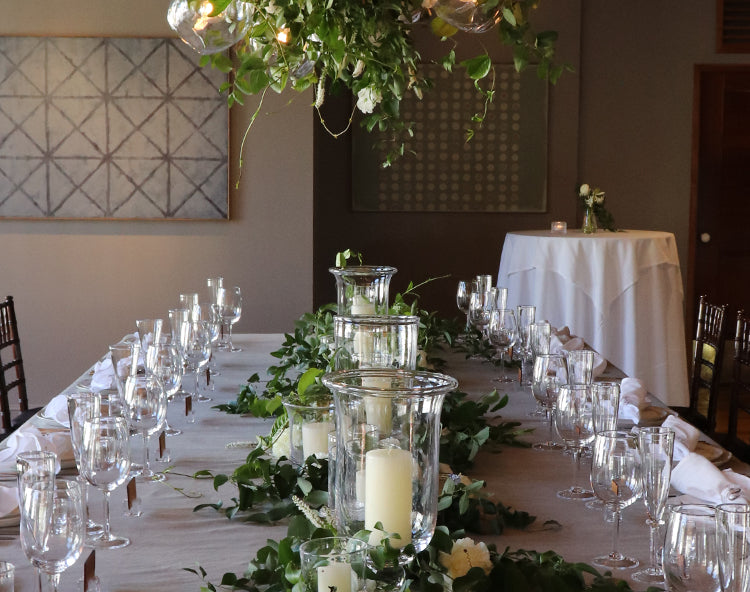 A long wedding reception table set with Simon Pearce glass hurricanes, pillar candles, and champagne flutes, decorated with a trailing greenery garland and hanging floral arrangement