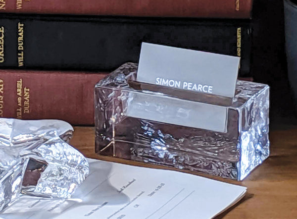 A Simon Pearce glass card holder displayed on a wooden desk beside books and wrapped silver tissue paper