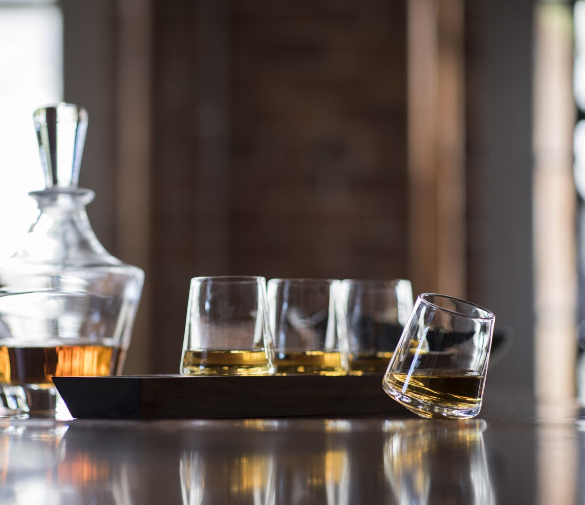Four clear glass whiskey glasses with amber liquid on a dark wood base, with a matching decanter in background.