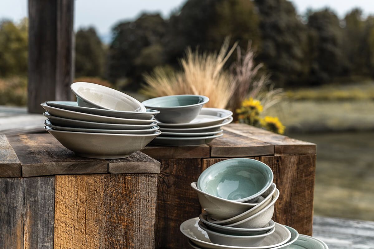 Stacked celadon and cream dinnerware bowls displayed on weathered wood surfaces outdoors.