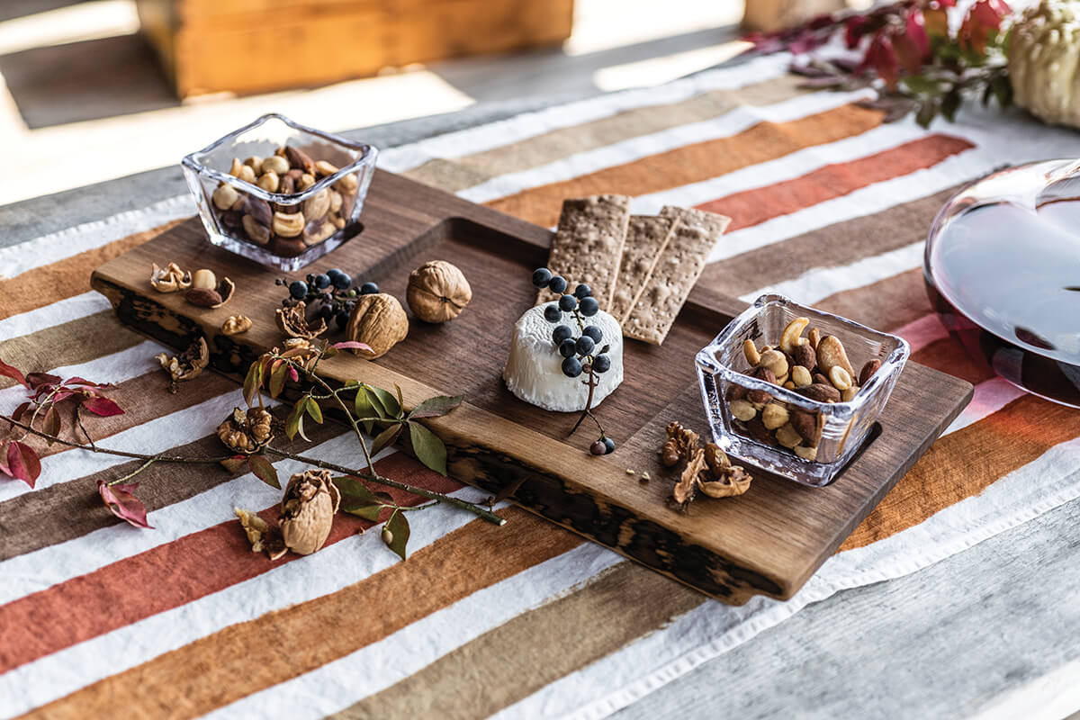 Black walnut board with two glass bowls filled with nuts, accompanied by dried botanicals and berries on striped runner.