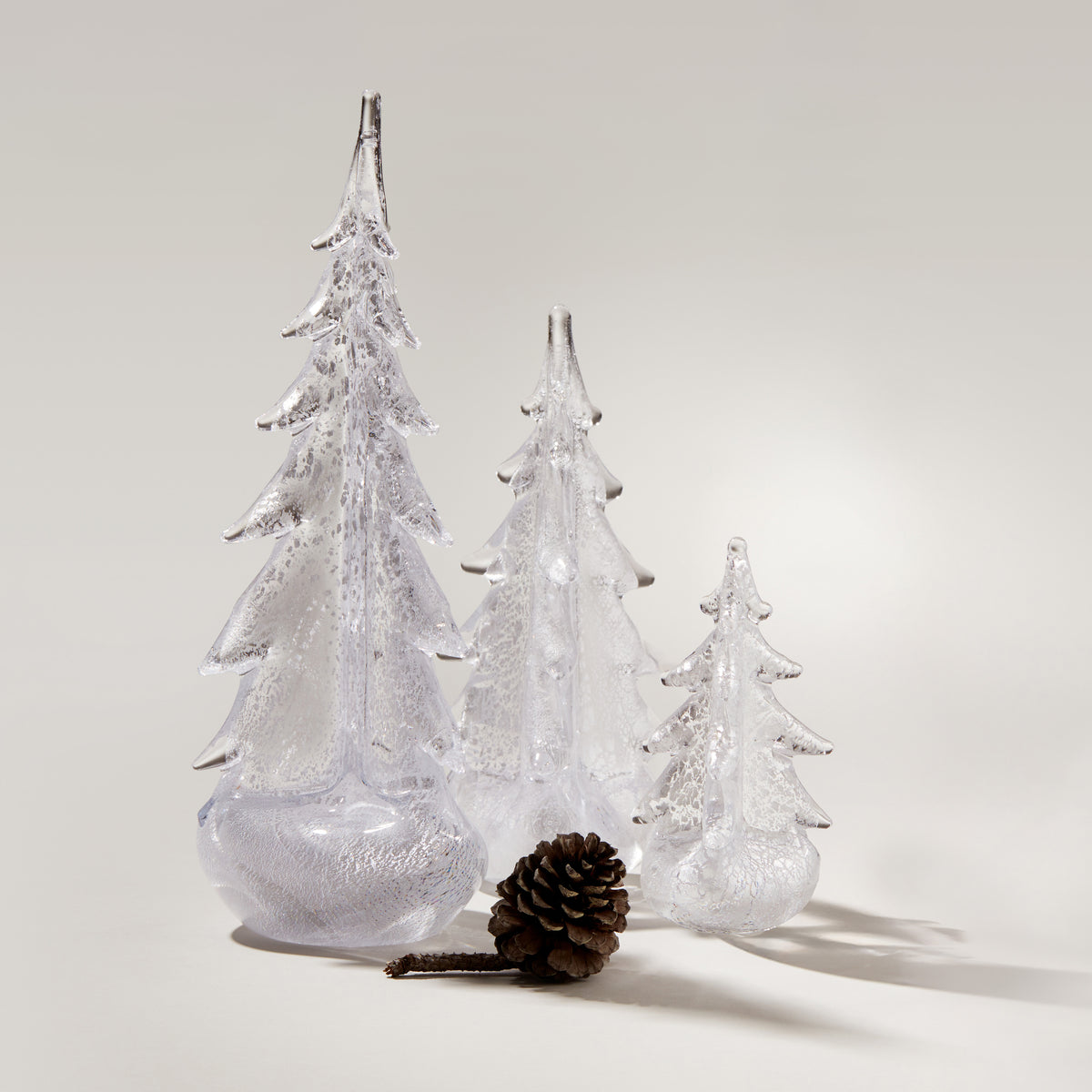 Three clear glass evergreen trees with frosted finish and a brown pinecone on neutral background.