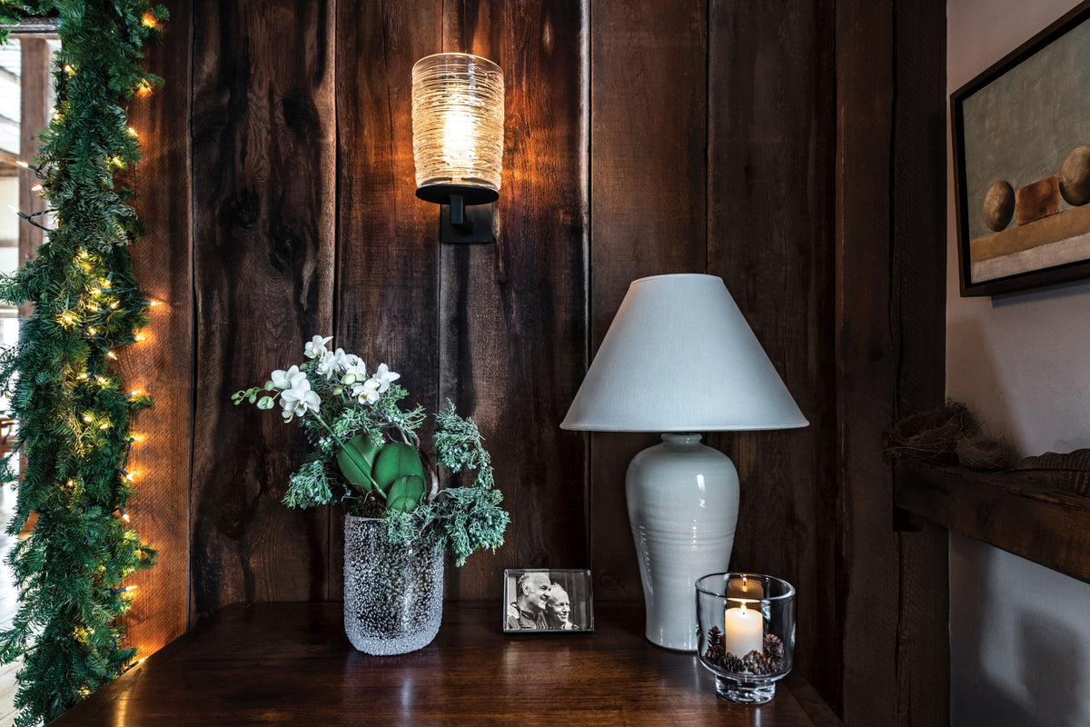Black metal wall sconce with ribbed glass cylinder shade, glowing warm light against dark wood paneling.
