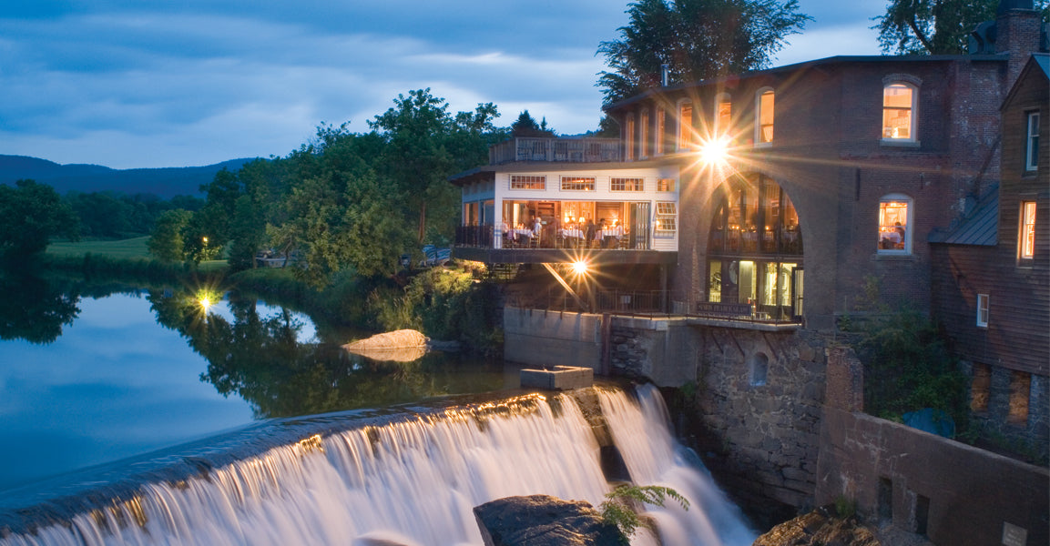 The Simon Pearce Mill in Quechee, Vermont at dusk, illuminated from within as guests dine on the terrace above the Ottauquechee River waterfall, with the historic brick mill building glowing against a blue twilight sky