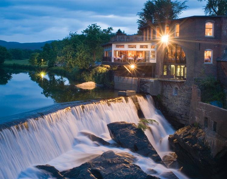 The Simon Pearce Mill in Quechee, Vermont at dusk, illuminated from within as guests dine on the terrace above the Ottauquechee River waterfall, with the historic brick mill building glowing against a blue twilight sky