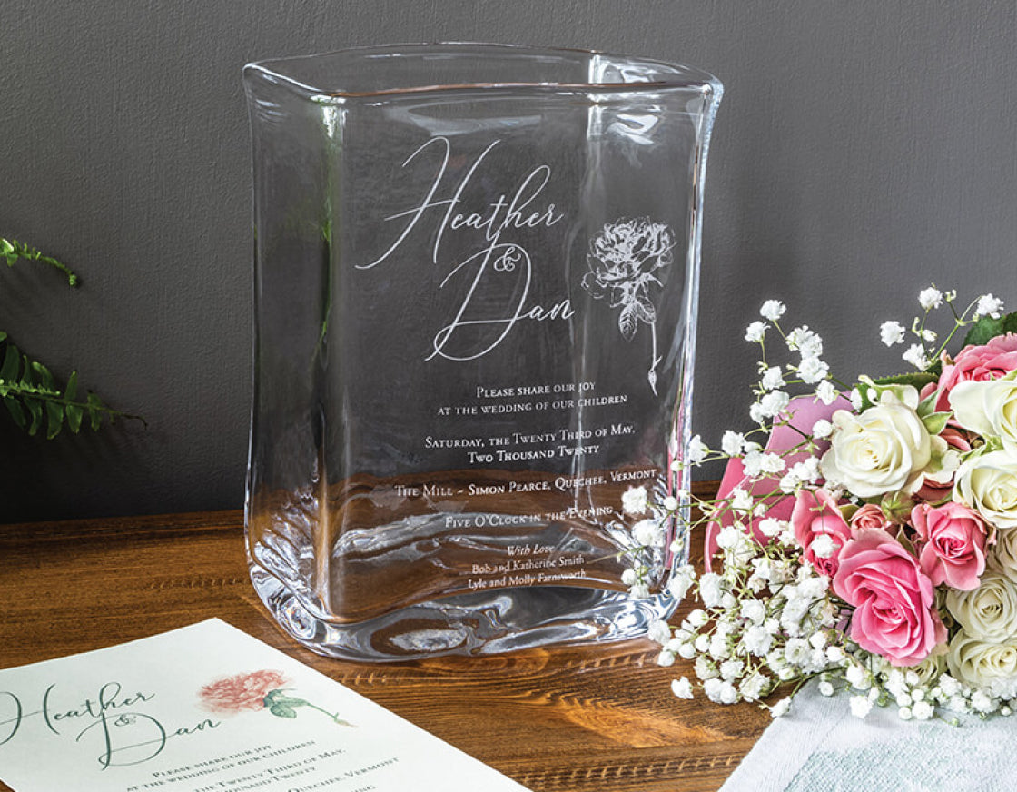 A large hand-blown glass Weston Vase engraved with a couple's wedding invitation, displayed on a wood surface beside pink and white roses and a matching paper invitation