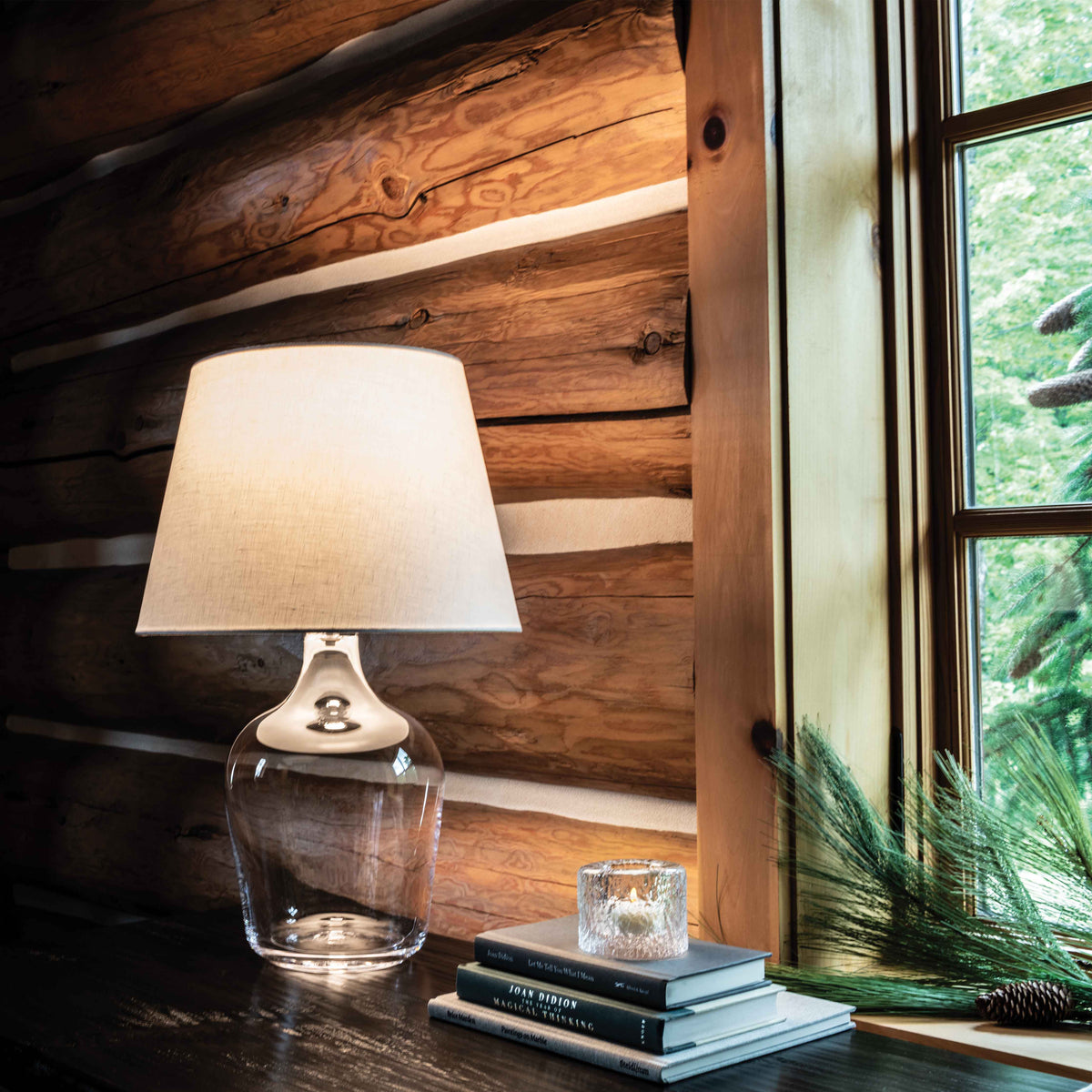 Table lamp with clear glass bottle base, cream fabric shade, and brushed metal neck on wooden log wall shelf.