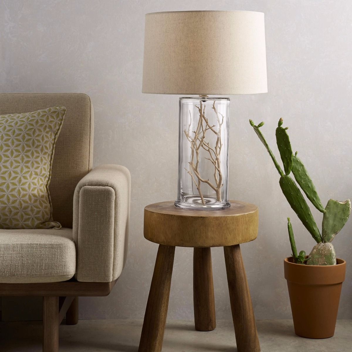 Cylindrical glass lamp base with dried branch filler and cream linen shade on wooden table.